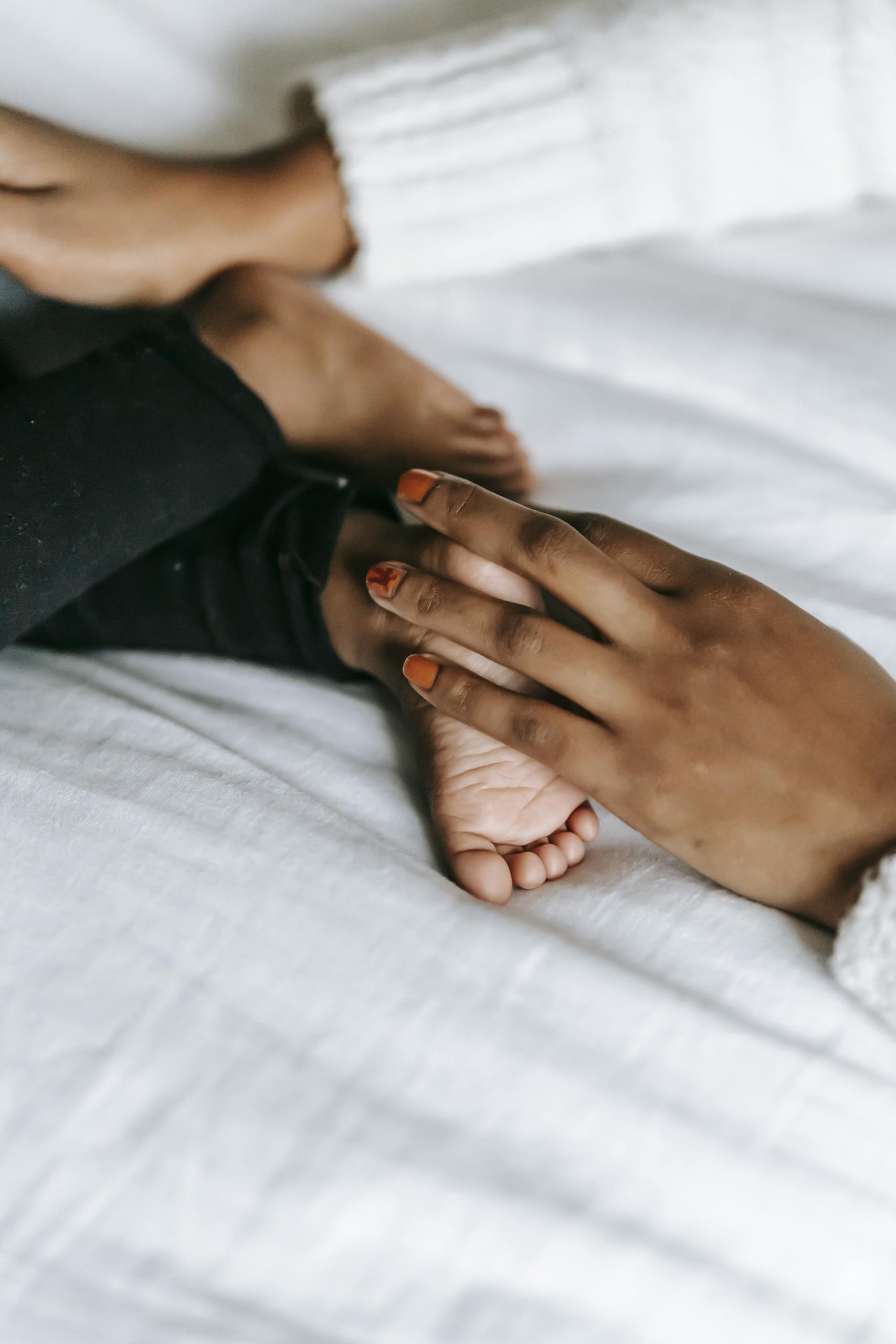 Unrecognizable African American mother touching barefoot foot of faceless infant baby while lying on white blanket in bedroom on bed